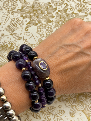 Three Amethyst Stack bracelets worn together on the wrist, featuring deep purple beads, gold-tone accents, and an oval centerpiece. ⠋